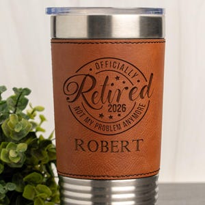May include: A stainless steel travel tumbler with a brown leather sleeve. The sleeve is engraved with the words "Officially Retired 2026 Not My Problem Anymore" and the name "Robert". The tumbler has a clear plastic lid.