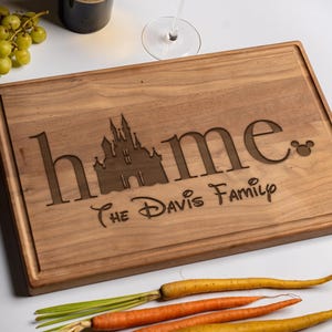 May include: A rectangular wooden cutting board with a carved design. The word "home" is displayed with a castle silhouette and a Mickey Mouse icon. The text "The Davis Family" is also carved into the board. A wine bottle, a glass of wine, and vegetables are in the background.