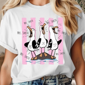 May include: A white short-sleeve t-shirt featuring a graphic of three black and white seagulls standing on a rock. The seagulls have orange beaks and feet. Behind them are vertical pink stripes. The text "Mrs. Smith" is printed multiple times.