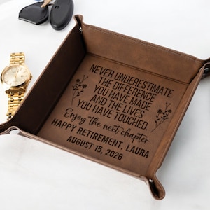 May include: A brown leather valet tray with a gold watch and keys. The tray has a personalized message: "NEVER UNDERESTIMATE THE DIFFERENCE YOU HAVE MADE... HAPPY RETIREMENT, LAURA AUGUST 15, 2026."