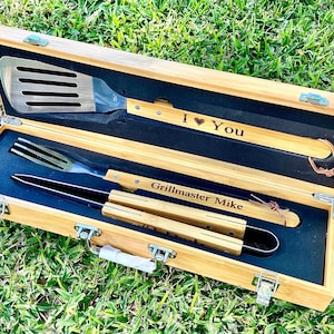 Puede incluir: Una caja de madera con un interior negro contiene un juego de herramientas para asar a la parrilla. Las herramientas incluyen una esp&aacute;tula, unas pinzas y un tenedor. La esp&aacute;tula y las pinzas tienen mangos de madera con el texto "Grillmaster Mike" y "I Love You" grabado en ellos.