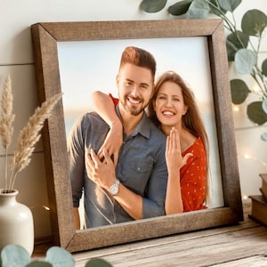 May include: A square, wooden picture frame with a photograph of a smiling couple. The frame is a warm brown color and rests on a wooden surface. Decorative elements include a vase with dried grass, greenery, and a candle.