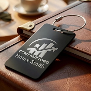 May include: A rectangular black luggage tag featuring a silver engraved graphic of a rising bar chart with an arrow, the text COMPANY LOGO, and the name Henry Smith. The tag includes a circular silver wire cable loop for secure attachment.