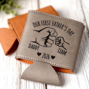 May include: Gray leatherette can cooler with the text "OUR FIRST FATHER'S DAY DADDY LIAM 2026" and a graphic of two fists bumping. The interior is orange, and the cooler is sitting on a white wooden surface.