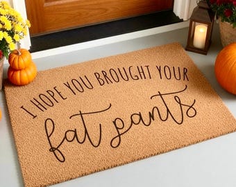 Puede incluir: Un felpudo rectangular de fibra de coco color canela con el texto impreso en negro I HOPE YOU BROUGHT YOUR fat pants. La alfombrilla tiene una textura fibrosa y gruesa, colocada sobre una superficie plana, mostrando un mensaje claro y legible.
