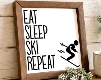 May include: A square wooden frame containing a white sign with black text that reads EAT SLEEP SKI REPEAT. To the right of the text is a black graphic of a person skiing. The frame is dark brown with a visible wood grain texture and finish.
