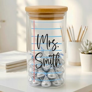 May include: A clear glass cylindrical jar with a light brown wooden lid. The jar features a printed design of blue horizontal lines and a red vertical margin line, resembling notebook paper. The black cursive text reads Mrs. Smith.