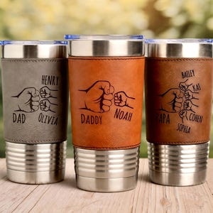 May include: Three personalized stainless steel travel mugs with leather sleeves. Each mug features a fist bump design with names like "Dad," "Henry," and "Olivia." The mugs have silver bases and lids, perfect for gifting.