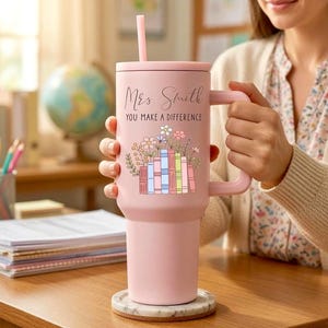 May include: A light pink tumbler with a handle and a straw, featuring the text "Mrs. Smith YOU MAKE A DIFFERENCE" above a graphic of colorful books topped with various flowers. The tumbler sits on a round, white marble-patterned coaster.