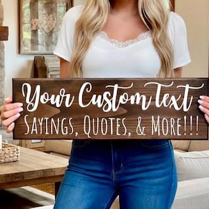 Peut inclure: Une enseigne en bois brun fonc&eacute; avec du texte blanc en cursive qui dit "Your Custom Text SAYINGS, QUOTES, & MORE!!!". L'enseigne rectangulaire est tenue par une personne portant un haut blanc et un jean bleu.