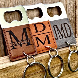 May include: Three personalized bottle openers with keychains. The openers have a silver metal top and a leatherette bottom. The leatherette bottoms are brown, tan, and gray, each with initials and a name engraved in black. The openers are on a wooden surface.