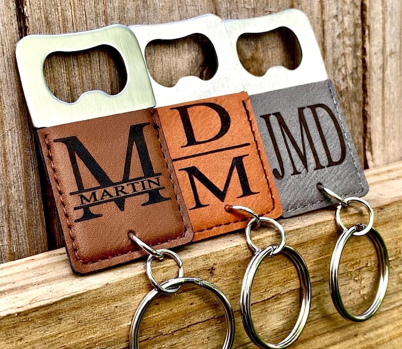 May include: Three personalized bottle openers with keychains. The openers have a silver metal top and a leatherette bottom. The leatherette bottoms are brown, tan, and gray, each with initials and a name engraved in black. The openers are on a wooden surface.