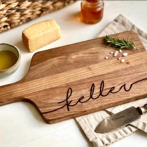 May include: A wooden cutting board with a handle, engraved with the name "Keller" in a cursive font. The board is surrounded by cheese, olive oil, rosemary, salt, and a knife. The board is approximately 12 inches long.