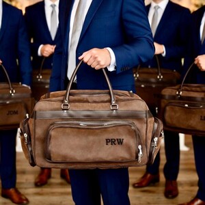 May include: Several men in navy blue suits hold brown leather duffel bags. The bags have a rectangular shape with a top handle and a front pocket. Each bag has initials embroidered on the front. The bags appear to be for a wedding or special event.