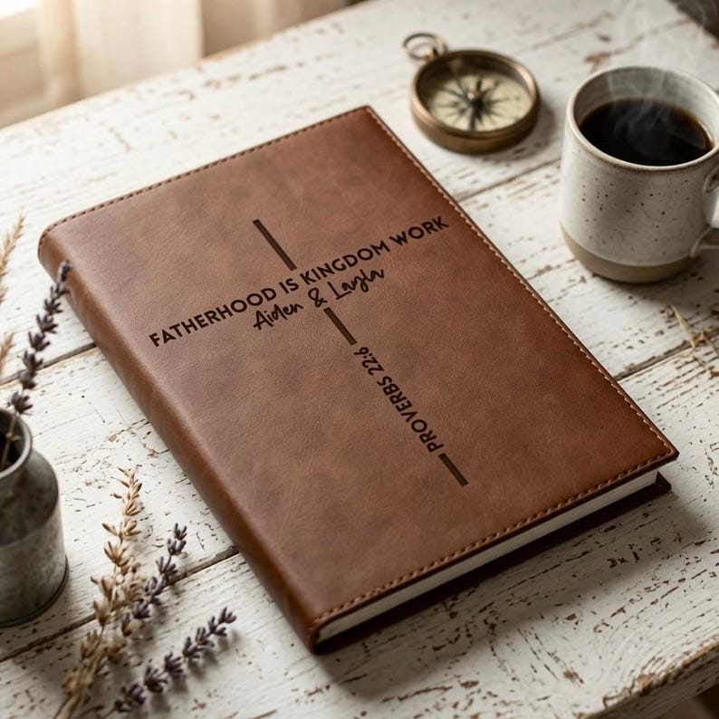 May include: A brown leather-like journal featuring a laser-engraved cross design. The text reads FATHERHOOD IS KINGDOM WORK, Aiden & Layla, and PROVERBS 22:6. The cover has visible stitching along the edges and a smooth, matte finish.