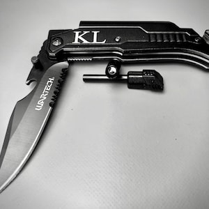 May include: A black multi-tool knife with a serrated blade, bottle opener, and a fire starter. The knife is open and the blade is visible. The knife has the text "WARTECH" and "KL" engraved on it.