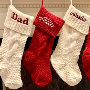 May include: Four cable-knit Christmas stockings hang from a fireplace mantel. Two are cream-colored with burgundy embroidered names, "Dad" and "Amelia." The other two are red with white embroidered names, "Allie" and another name.