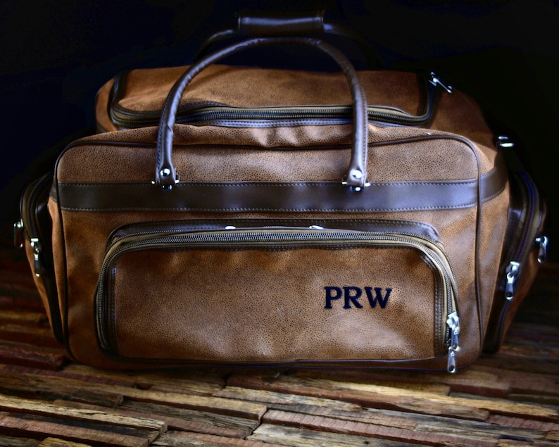 May include: Brown duffel bag with a textured finish and dark brown leather accents. Features a top handle, multiple zippered compartments, and the initials "PRW" embroidered on the front.