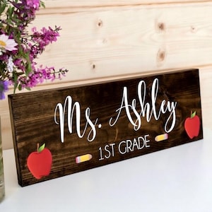 May include: A dark stained wooden sign with the name "Ms. Ashley" in white script. The sign also includes "1st Grade" in white text, with red apple and pencil graphics. The sign is a rectangular shape.