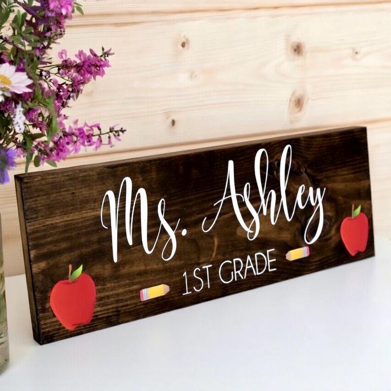 May include: A dark stained wooden sign with the name "Ms. Ashley" in white script. The sign also includes "1st Grade" in white text, with red apple and pencil graphics. The sign is a rectangular shape.