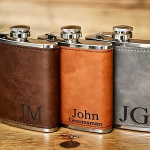 May include: Three personalized flasks in brown, orange, and gray leather. Each flask has a silver cap and trim. The brown flask has the initials "JM", the orange flask says "John Groomsman", and the gray flask has the initials "JG".