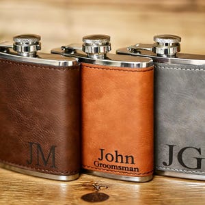 May include: Three personalized flasks in brown, orange, and gray leather. Each flask has a silver cap and trim. The brown flask has the initials "JM", the orange flask says "John Groomsman", and the gray flask has the initials "JG".