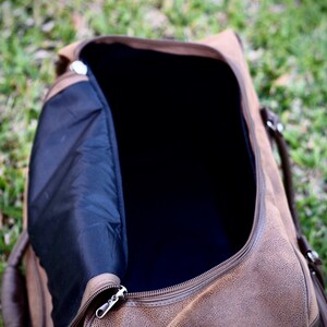 May include: Open brown duffel bag with a black interior and a silver zipper. The bag has brown handles and is sitting on a bed of green grass. The bag is made of a textured material.