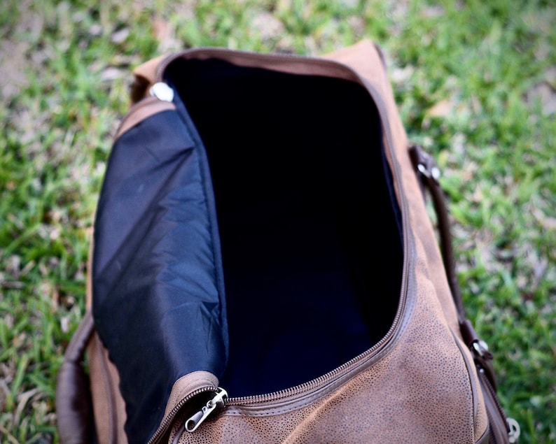 May include: Open brown duffel bag with a black interior and a silver zipper. The bag has brown handles and is sitting on a bed of green grass. The bag is made of a textured material.