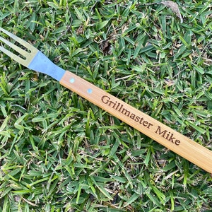 Puede incluir: Un tenedor para asar con mango de madera y p&uacute;as de acero inoxidable. El mango est&aacute; grabado con el texto "Grillmaster Mike".