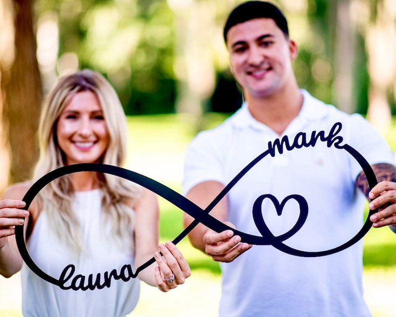 May include: A couple hold a black infinity symbol with the names "Laura" and "mark" written on it. The symbol also includes a heart in the centre.