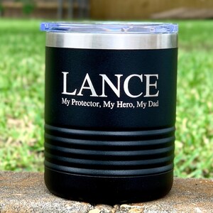 May include: Black stainless steel tumbler with a silver lid. The tumbler has the name "LANCE" engraved on the front, along with the text "My Protector, My Hero, My Dad".