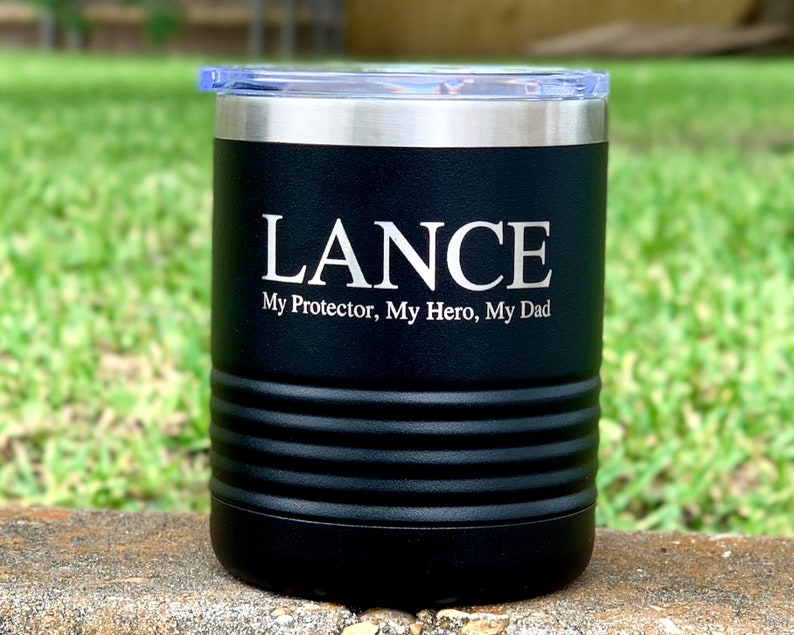 May include: Black stainless steel tumbler with a silver lid. The tumbler has the name "LANCE" engraved on the front, along with the text "My Protector, My Hero, My Dad".