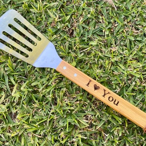 Puede incluir: Una esp&aacute;tula de madera con una hoja de acero inoxidable y un mango que dice "Te amo" con un s&iacute;mbolo de coraz&oacute;n. La esp&aacute;tula est&aacute; sobre un lecho de hierba verde.