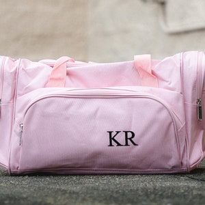 May include: A light pink duffel bag with a front zippered pocket and side pockets. The bag has a top handle and a shoulder strap. The initials "KR" are embroidered on the front of the bag.