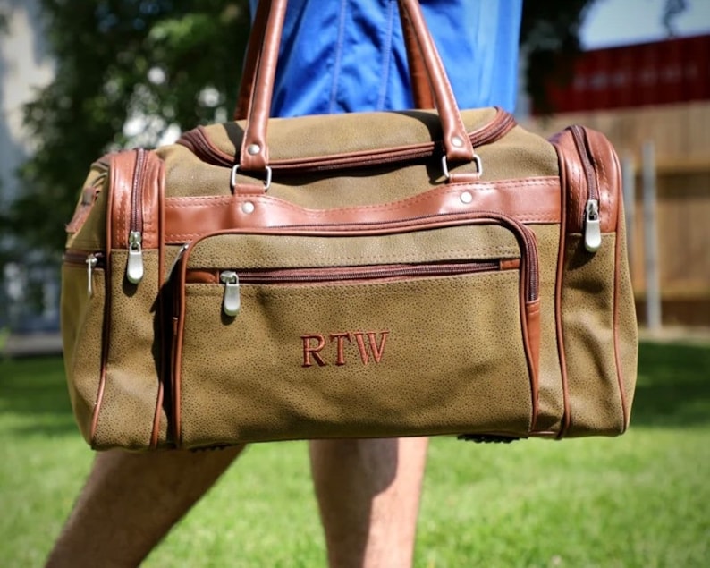 May include: A brown and tan duffel bag with brown leather accents. The bag has a front pocket, side pockets, and a top handle. The initials "RTW" are embroidered on the front. The bag has silver zippers and a shoulder strap.