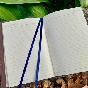 May include: An open notebook with cream-colored lined pages featuring a date header and a day-of-the-week selection box. Two dark blue ribbon bookmarks are attached to the spine, resting across the pages. The notebook has a dark brown cover.