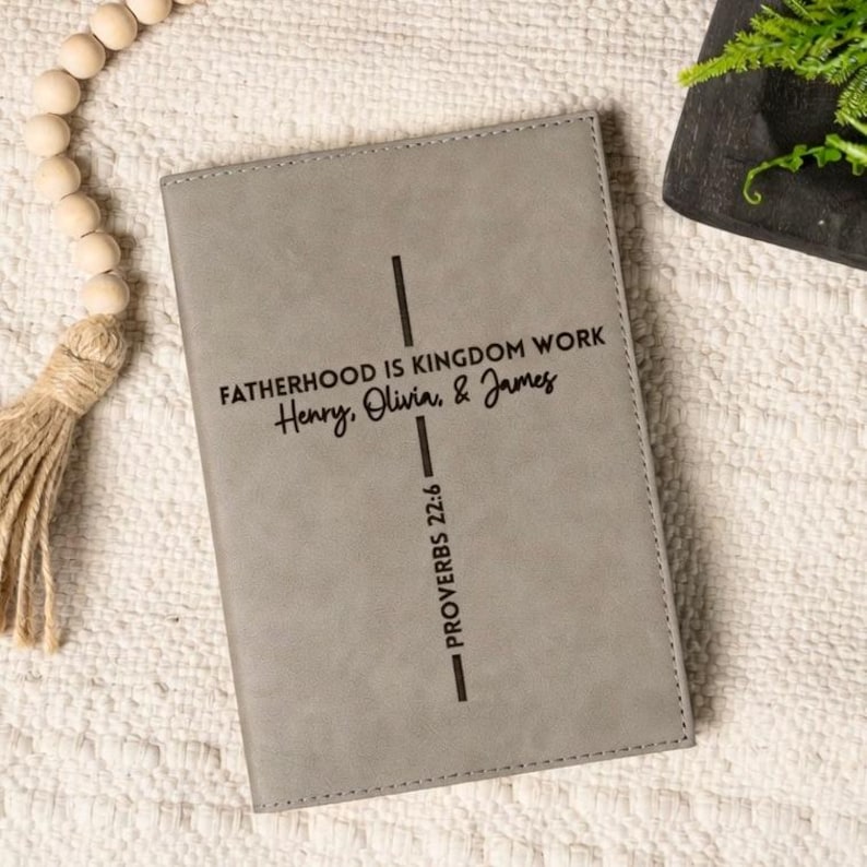 May include: A rectangular gray notebook with a vertical black cross design. The text reads FATHERHOOD IS KINGDOM WORK, Henry, Olivia, & James, and PROVERBS 22:6. The cover has a smooth, matte finish with stitched edges along the perimeter.