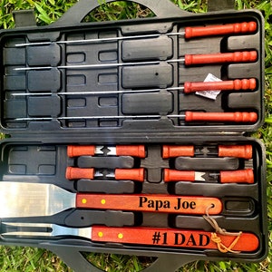 Puede incluir: Una maleta de transporte negra con un asa roja contiene un juego de herramientas para asar a la parrilla. Las herramientas incluyen pinchos, una esp&aacute;tula y un tenedor. La maleta tiene un asa de madera con el texto "Papa Joe #1 Dad" grabado en ella.