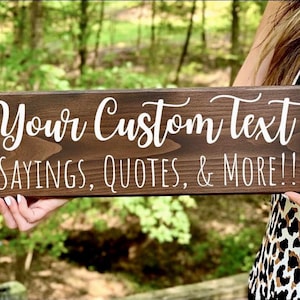 Peut inclure: Une enseigne rectangulaire en bois brun fonc&eacute; avec du texte blanc en cursive qui dit "Your Custom Text SAYINGS, QUOTES, & MORE!!!". L'enseigne est tenue devant un arri&egrave;re-plan vert flou.