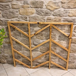 Puede incluir: Un enrejado de madera con un diseño geométrico. El enrejado está hecho de madera de color marrón claro y presenta una serie de soportes angulados, creando un patrón en zigzag. El enrejado es independiente y se apoya contra una pared de piedra.