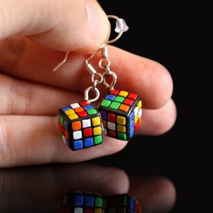 May include: A pair of silver earrings with colorful cube-shaped charms. The charms are reminiscent of a Rubik's Cube and feature a variety of colors, including red, blue, green, yellow, orange, and white.