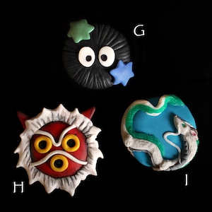 May include: Four handmade clay charms inspired by Studio Ghibli films. The charms feature a black and white No Face character, a red and white mask, a blue and green No Face character, and a white and green Totoro character.