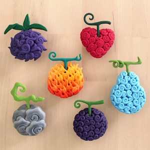 May include: Six colorful clay sculptures of devil fruits from the anime One Piece. The fruits are shaped like a heart, a pear, a cloud, and a flame. Each fruit has a unique swirl pattern and a green stem.