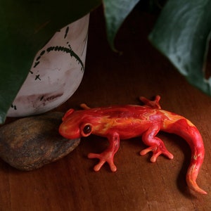 Puede incluir: Una figurita de salamandra de color naranja y amarillo brillante hecha de arcilla polimérica. La salamandra está posada sobre su vientre con las patas extendidas y la cola enroscada.
