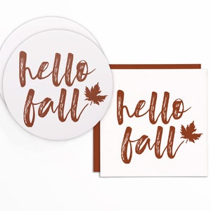May include: White round coasters and a square card with the words "hello fall" in brown script and a brown maple leaf. The coasters are stacked, and the card has a brown border.