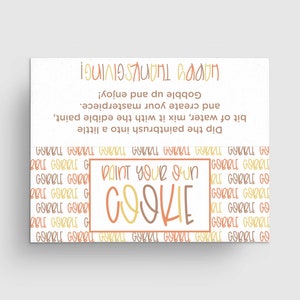 May include: A white card with the text "Paint Your Own Cookie" in orange and yellow lettering. The card has a background of repeating "Gobble Gobble" text in orange, yellow, and brown.