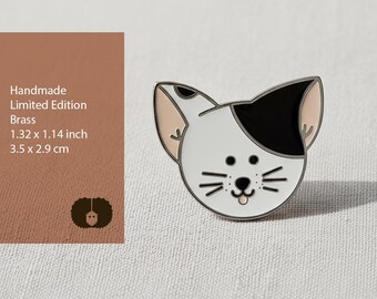 Könnte beinhalten: Handgefertigte, limitierte Katzenbrosche aus Messing. Die Brosche zeigt ein weißes Gesicht mit einem schwarzen Fleck über einem Auge, schwarzen Schnurrhaaren und einer rosa Zunge. Misst 3,5 x 2,9 cm.