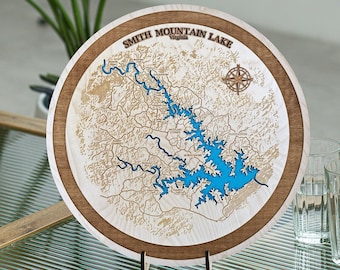 Puede incluir: Una placa redonda de madera con un mapa del lago Smith Mountain, Virginia. El lago está delineado en azul y el área circundante está grabada con líneas marrones. Una brújula se encuentra en la esquina superior derecha.