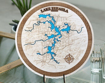 May include: A wooden round plaque with a map of Lake Eufaula, Oklahoma. The lake is highlighted in blue and the surrounding area is engraved in brown. The plaque has a compass rose in the lower right corner and the words "Lake Eufaula, Oklahoma" at the top.