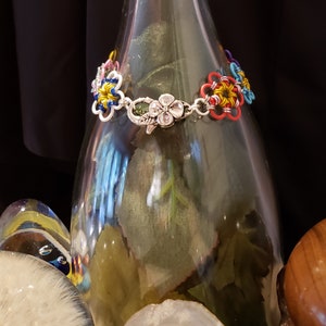 以下が含まれることがあります： 銀色の留め具が付いたカラフルな花の形をしたブレスレット。ブレスレットは、小さなカラフルなビーズでできており、銀色のチェーンに通されています。